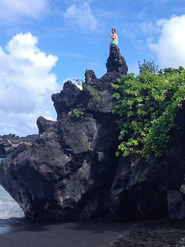 A woman stands atop a jagged volcanic rock formation at a black sand beach in Maui, Hawaii, surrounded by lush greenery.