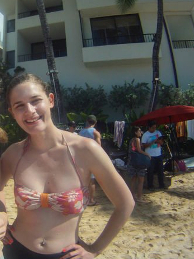 A smiling woman in a floral bikini stands on a sandy beach in Hawaii, with a hotel, palm trees, and beachgoers in the background.