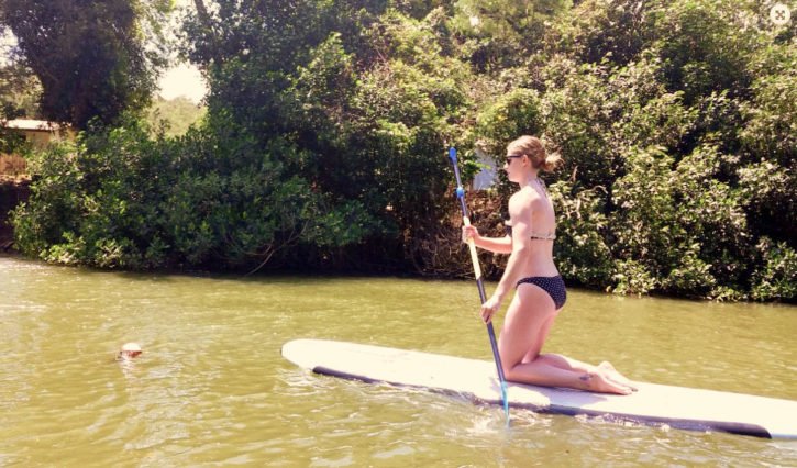 A woman in a black polka-dot bikini kneels on a paddleboard, holding a paddle while navigating a calm river in Hawaii, surrounded by lush greenery.