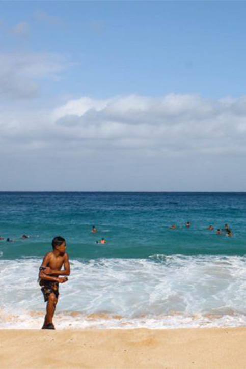 A young boy playing on the sandy shore of a Hawaiian beach while people swim in the deep blue ocean under a partly cloudy sky.