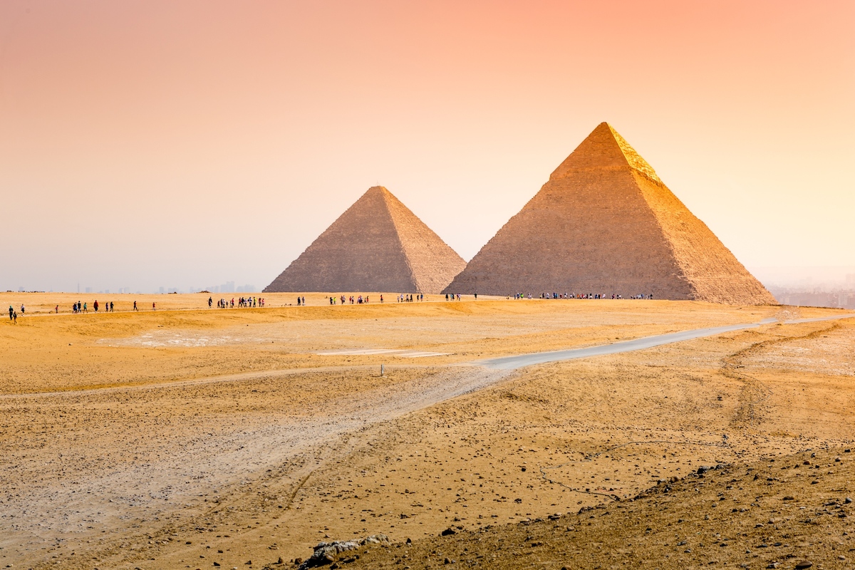 The Great Pyramids of Giza in Egypt standing majestically under a warm, orange-hued sunset sky with scattered tourists nearby.