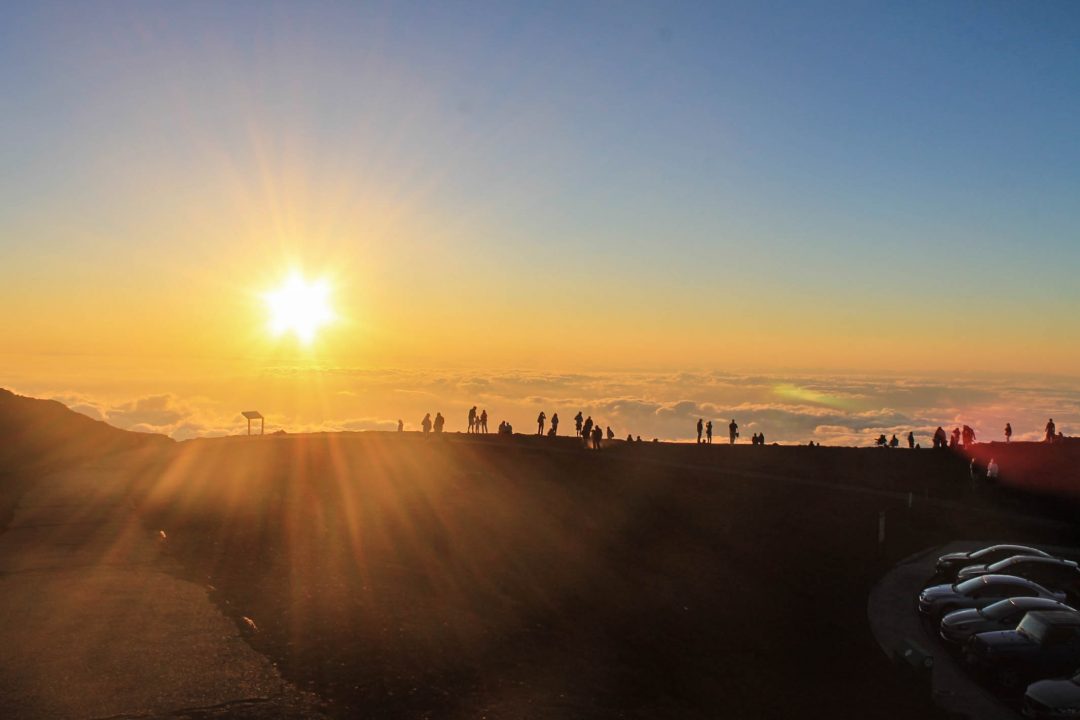 A group of people stands silhouetted against a golden sunrise above the clouds at the Haleakalā summit in Maui, Hawaii.