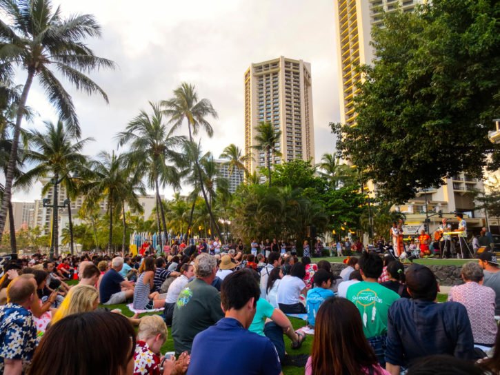 A crowd gathers on the grass to watch a live outdoor performance at Waikiki Beach in Honolulu, Hawaii, surrounded by palm trees and tall buildings.